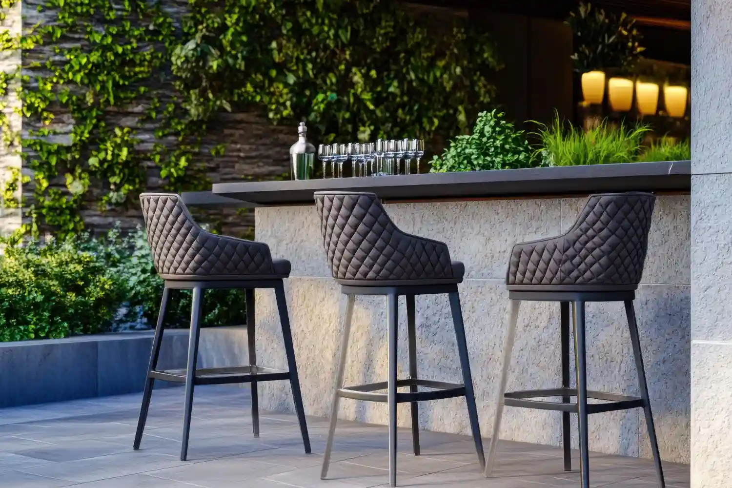 Three black outdoor bar stools with quilted seats and metal footrest bars at a stone-clad garden bar counter