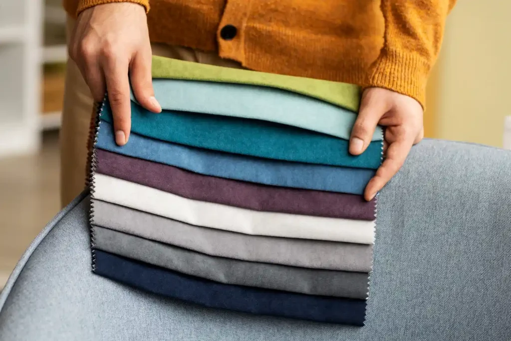 Hand holding a stack of contract restaurant chair fabric swatches in teal, green, grey and navy over an upholstered chair arm