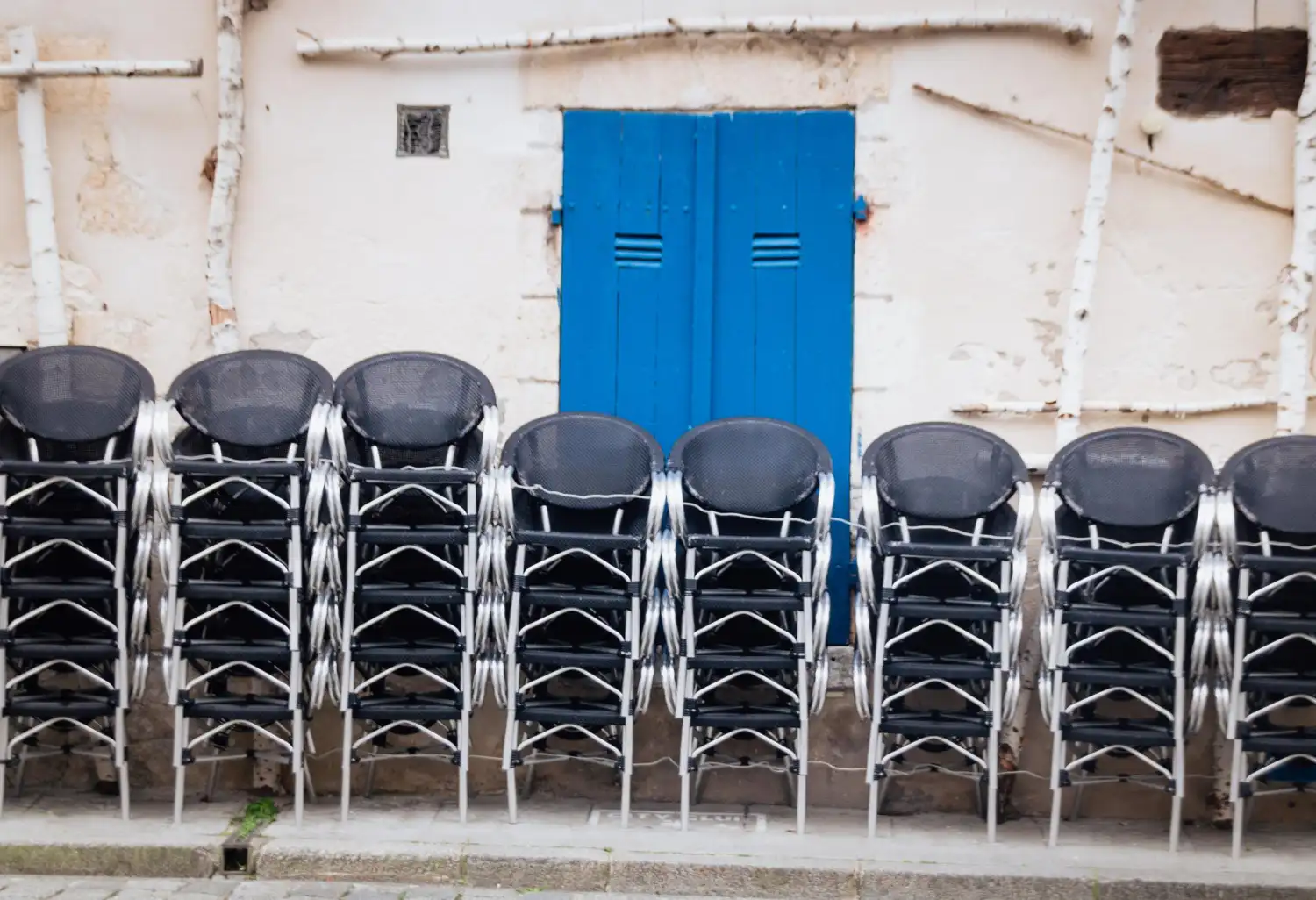 Rows of plain black metal restaurant chairs stacked against a whitewashed wall beside a blue door