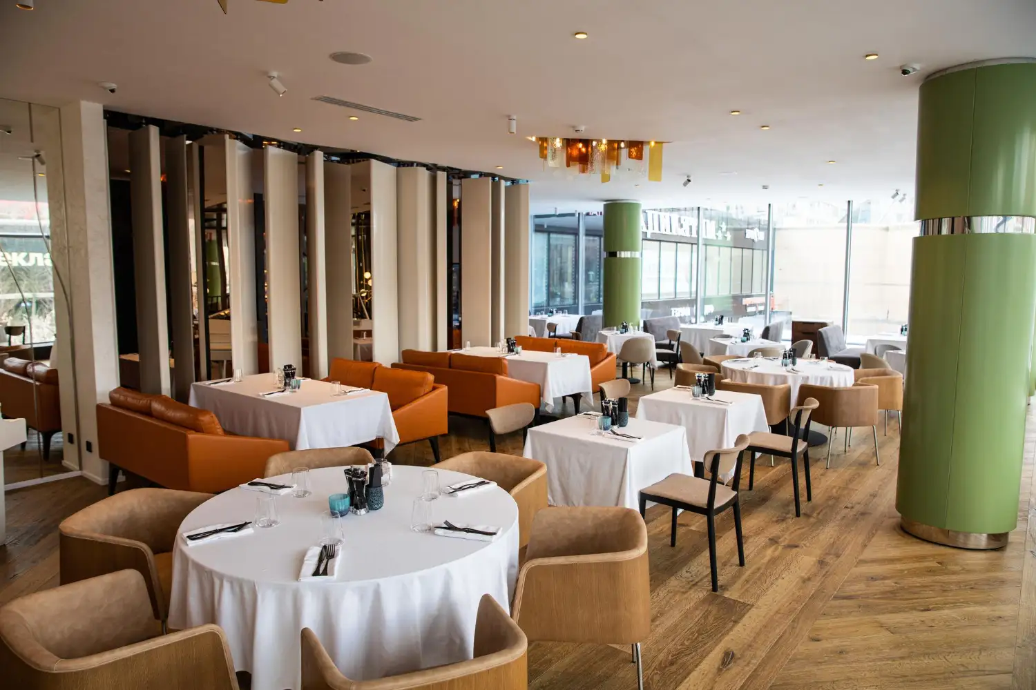 Large hotel restaurant with round linen-covered tables, tan upholstered armchairs and banquette seating by full-height glazing