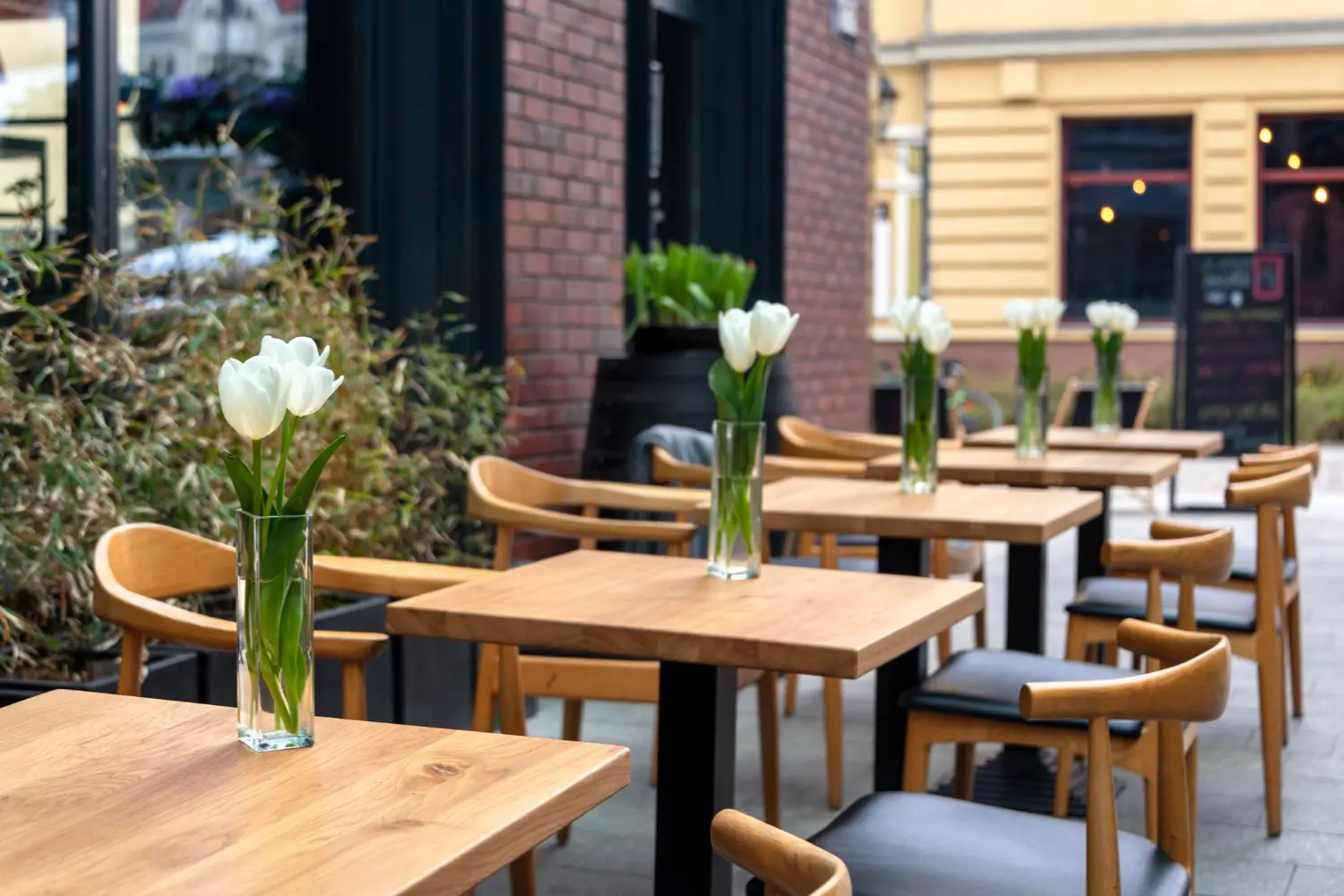 Solid timber terrace tables with light timber armchairs and flowers, set outside a restaurant on a city street