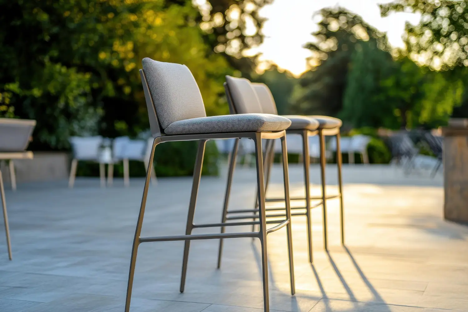Pale upholstered outdoor bar stools on slim metal legs with footrest rings, on a stone terrace at sunset